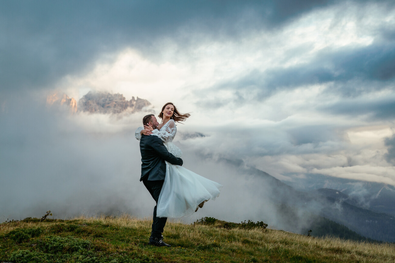 A man in a suit lifts a woman in a white dress on a grassy hilltop, surrounded by clouds and misty mountains in the background.