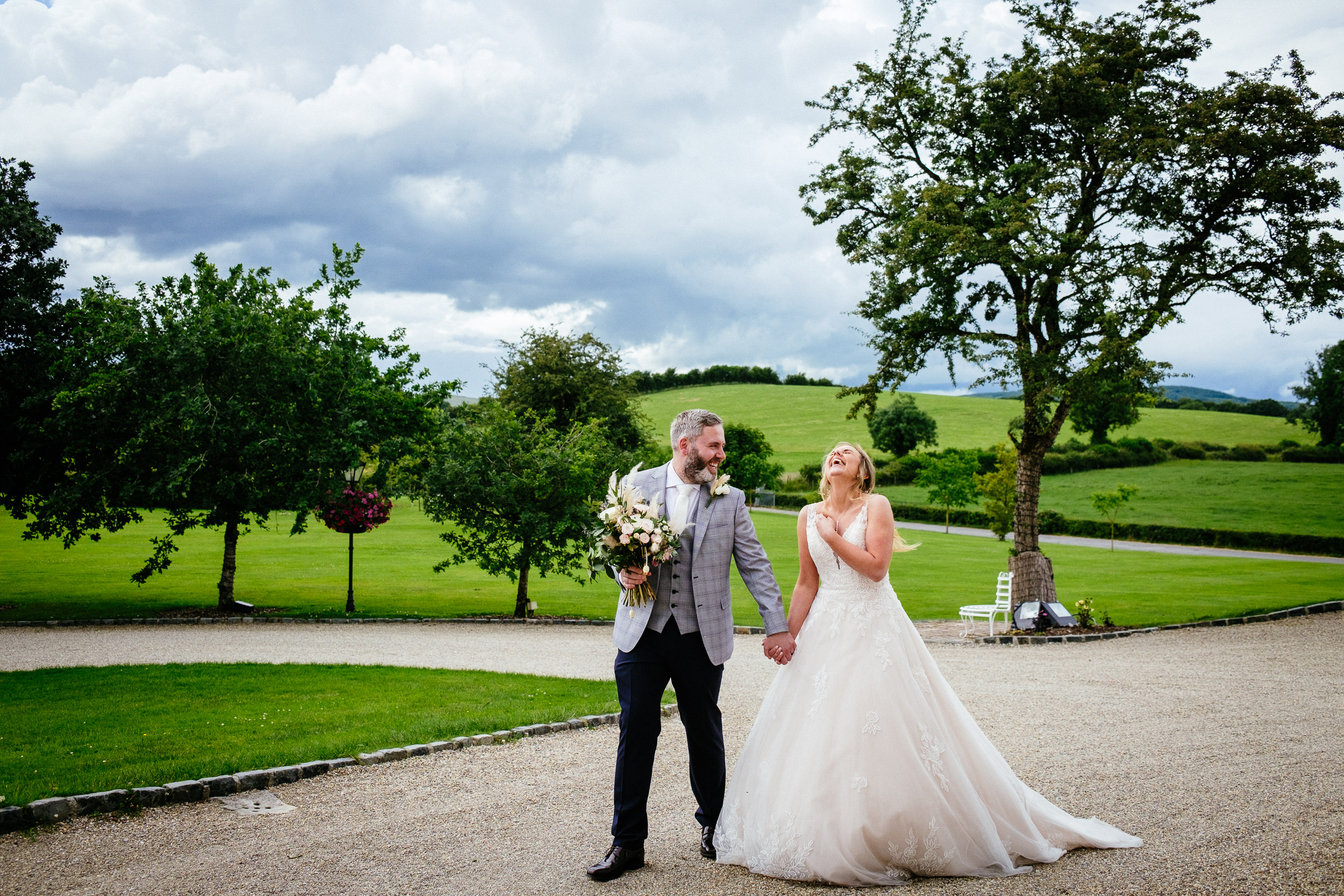 A couple in wedding attire holding hands, with a bouquet, walking on a gravel path surrounded by green lawns and trees under a cloudy sky whilst Love at Clonabreany House.