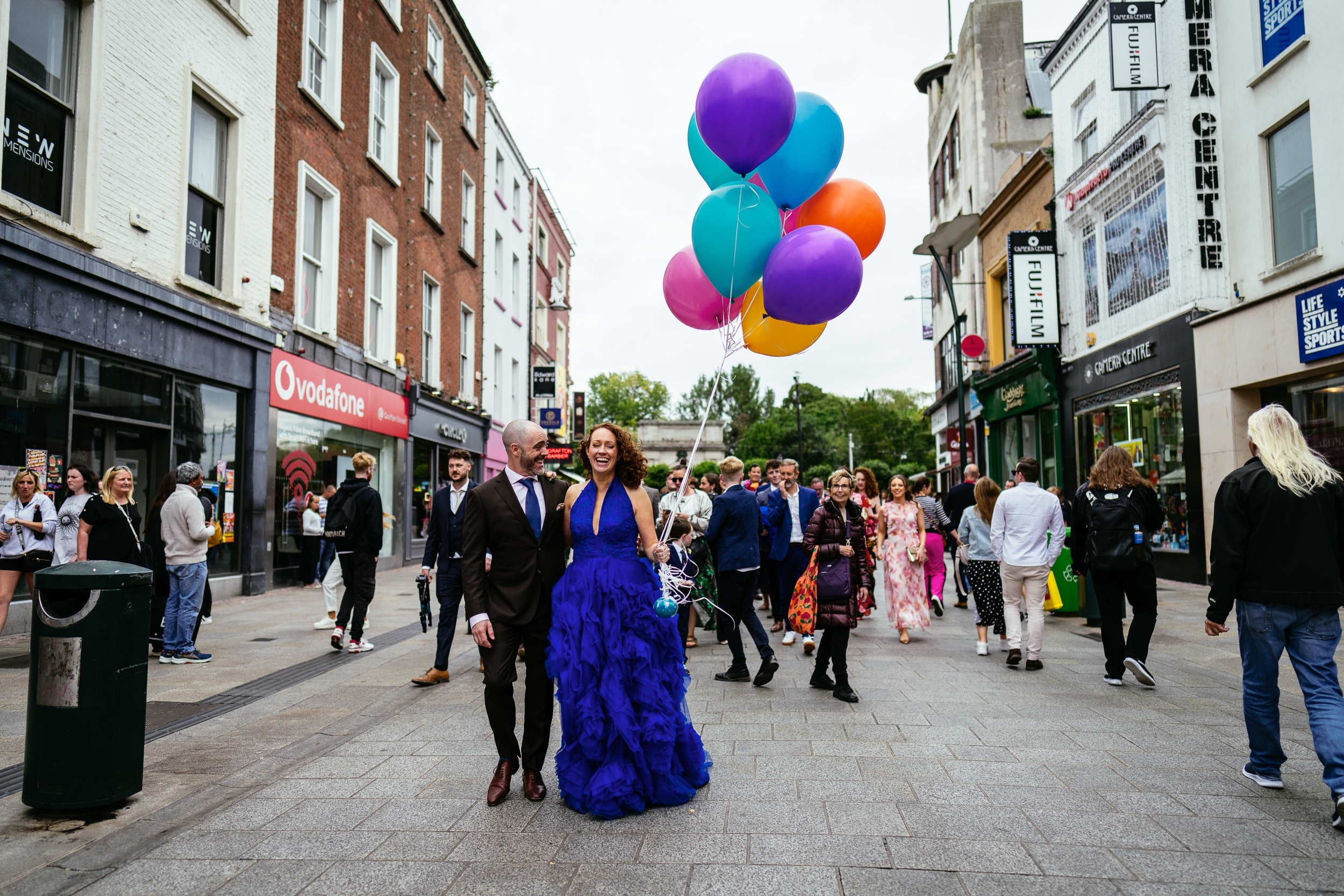 A couple walks down a bustling street with colorful balloons. The person on the left wears a suit, and the person on the right is in a bright blue dress. Passersby are visible in the background.