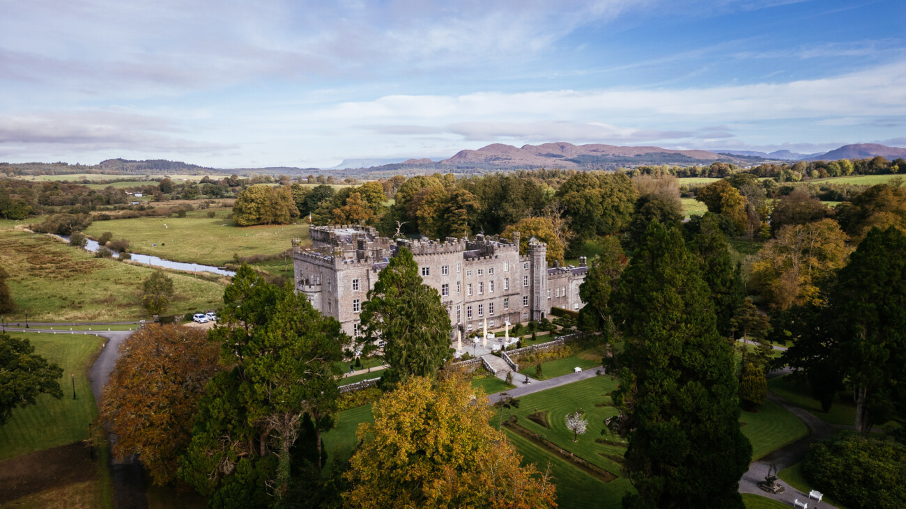 Aerial view of an old stone castle surrounded by trees and rolling hills under a cloudy sky.