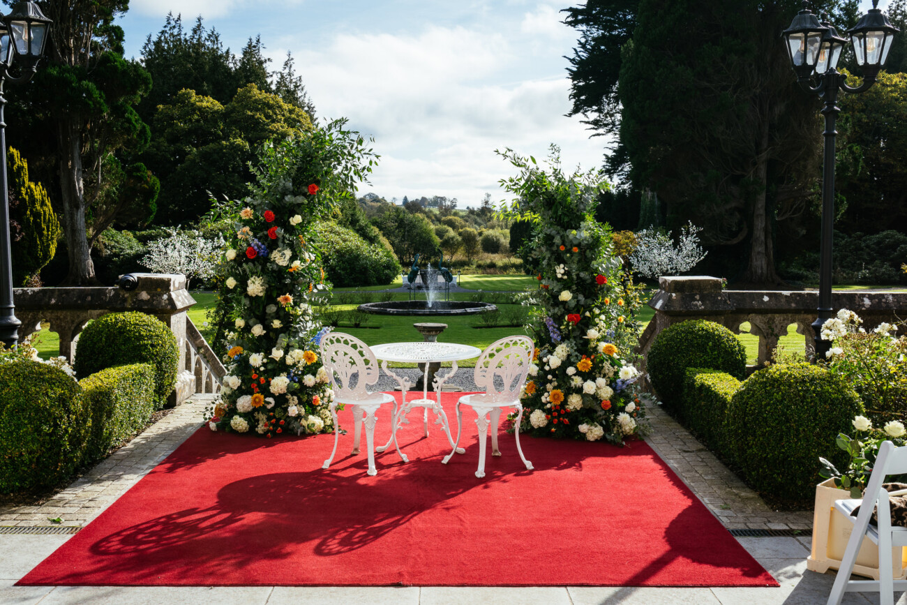 A scenic garden with a red carpet and a white metal table with two chairs. Floral arrangements flank the setup, and a fountain is visible in the background.
