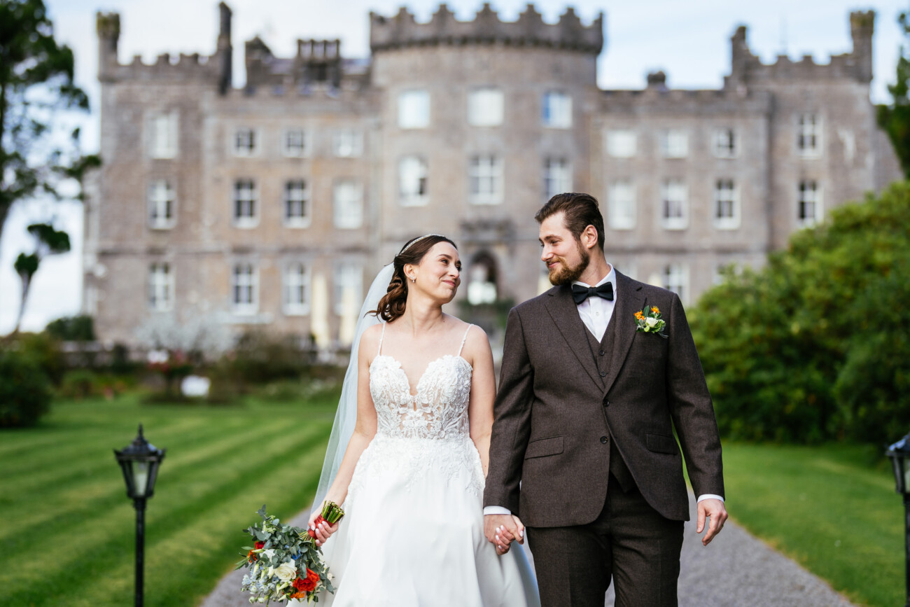 A bride and groom hold hands, walking in front of a stone castle. The bride wears a white gown and holds a bouquet; the groom wears a brown suit with a bow tie. Trees and grass line the pathway.