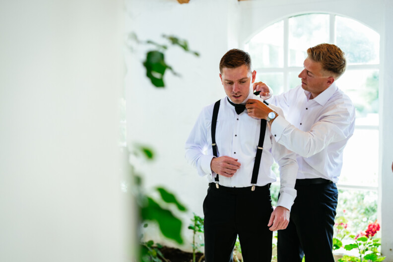 Two men in white shirts are in a room with a large window. One adjusts the other's bow tie. Red flowers and greenery are in the foreground.