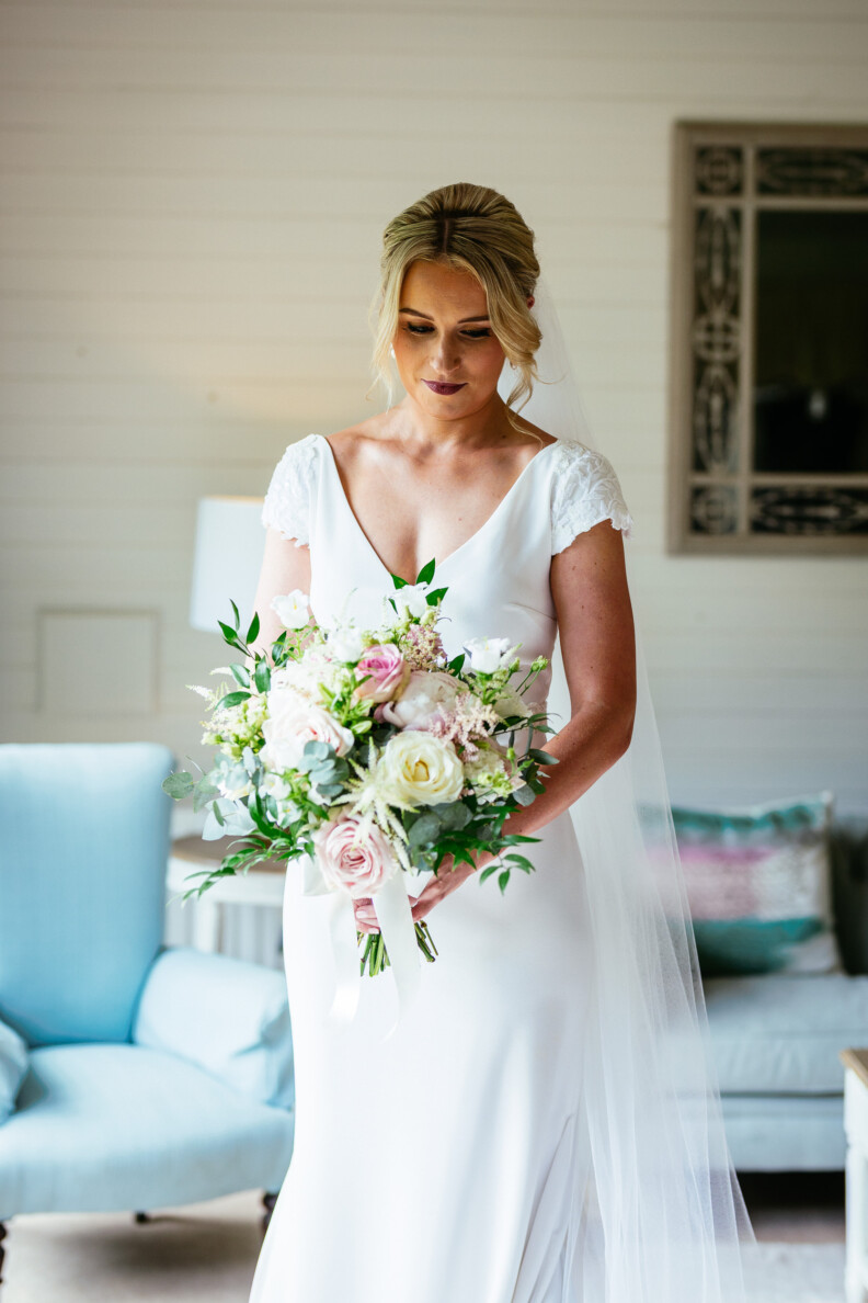 A bride in a white dress holds a bouquet of flowers. She stands in a room with a light blue armchair and a decorative mirror on the wall.