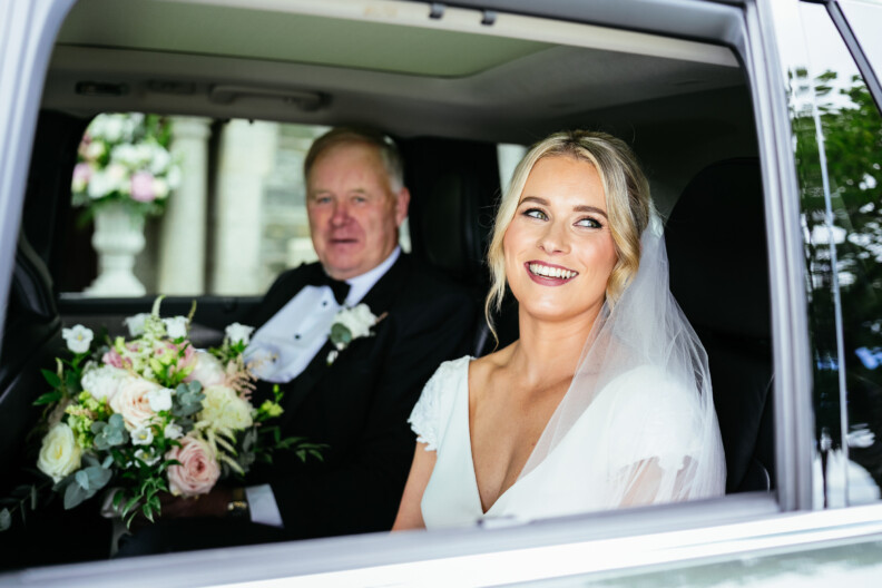 A bride in a white dress and veil sits in a car holding a bouquet, alongside an older man in formal attire.