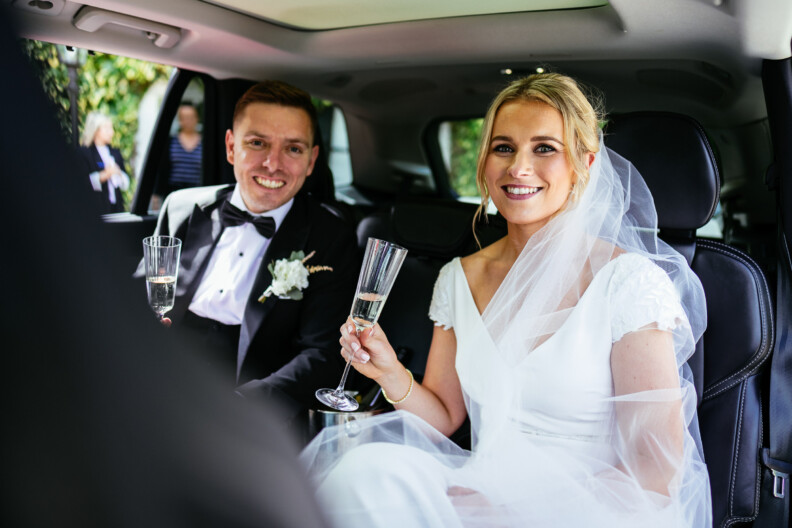 A bride and groom sit in a car, smiling and holding champagne glasses. The bride wears a white dress and veil, while the groom wears a black tuxedo with a bow tie.