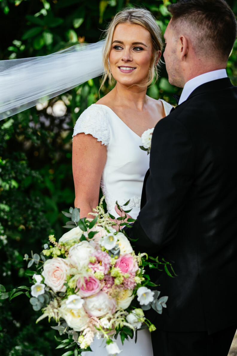 A bride and groom stand together outdoors, with the bride holding a bouquet of white and pink flowers. The groom faces away from the camera.