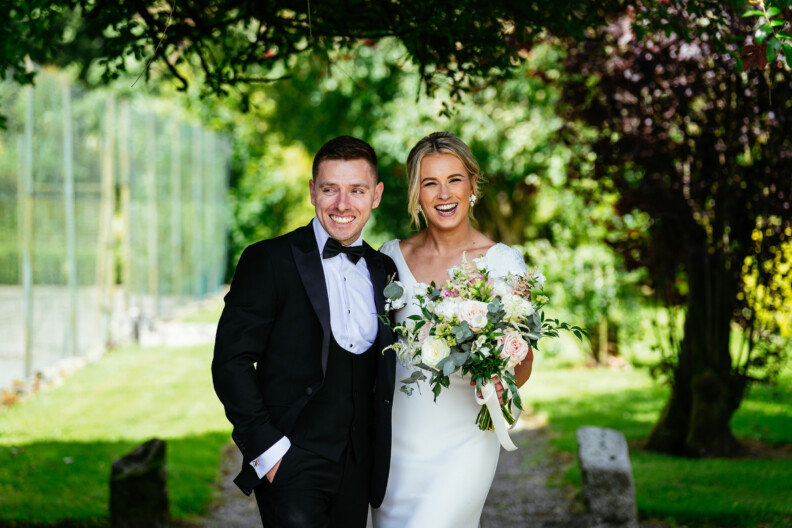 A smiling couple in formal wear, with the man in a black tuxedo and the woman in a white dress holding a bouquet, stand outdoors under greenery.
