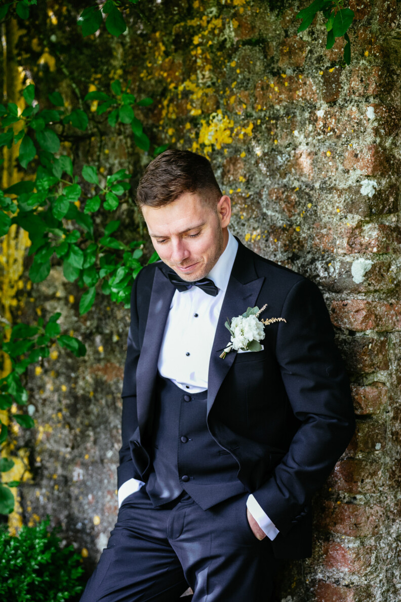 Man in a black tuxedo with a white boutonniere leans against a moss-covered brick wall, looking down.