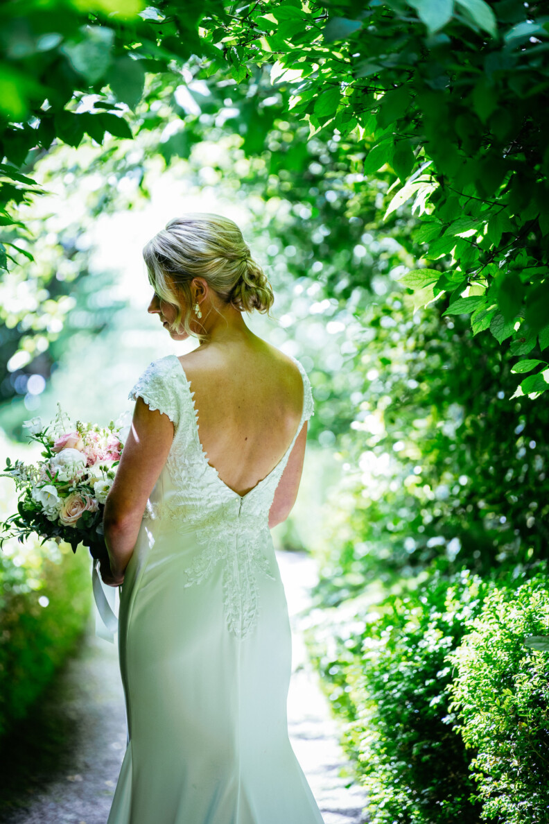 Bride in a white, lace-back dress holds a bouquet, standing on a garden path surrounded by lush greenery.