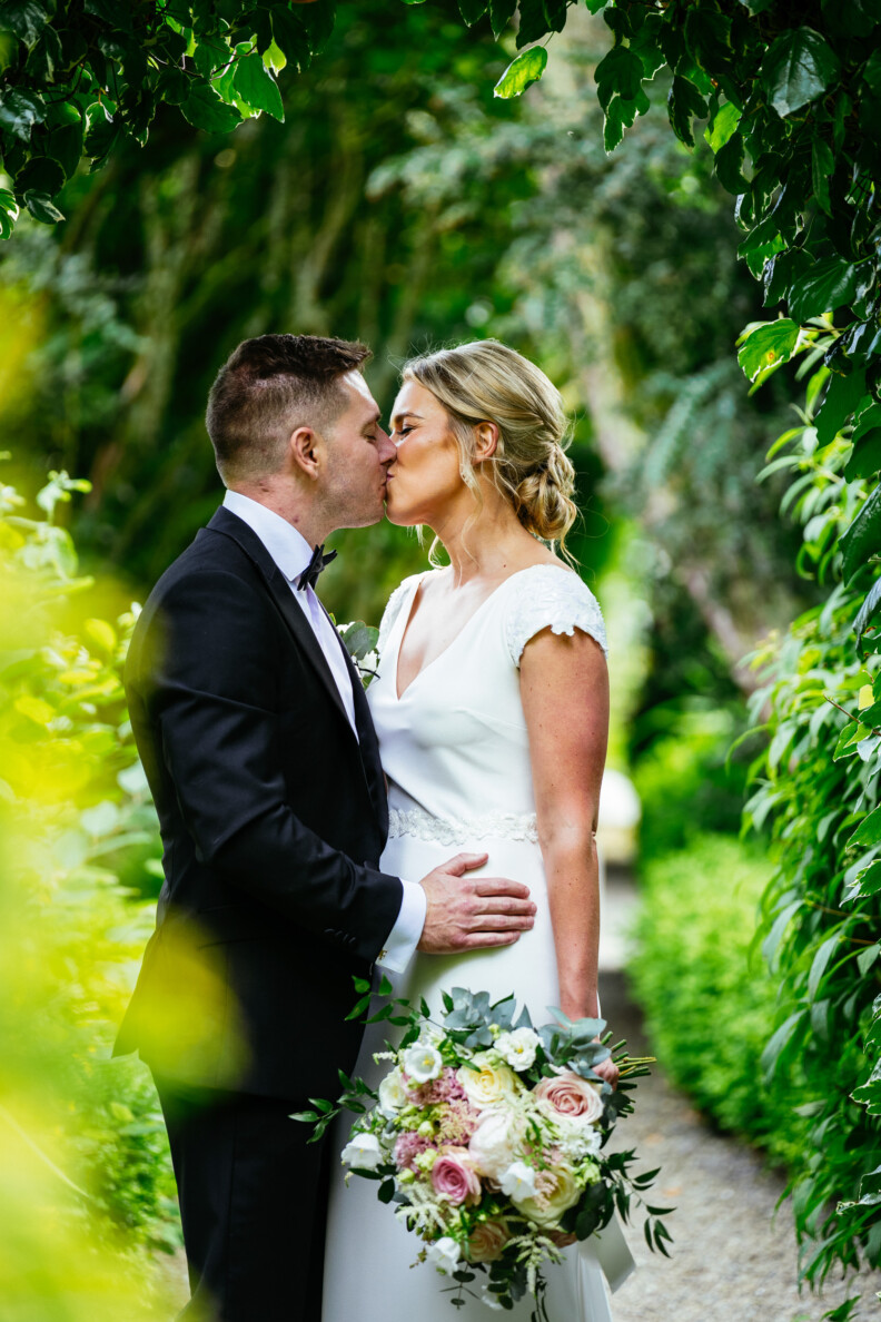 A bride and groom kiss outdoors, surrounded by greenery. The bride holds a bouquet of flowers.