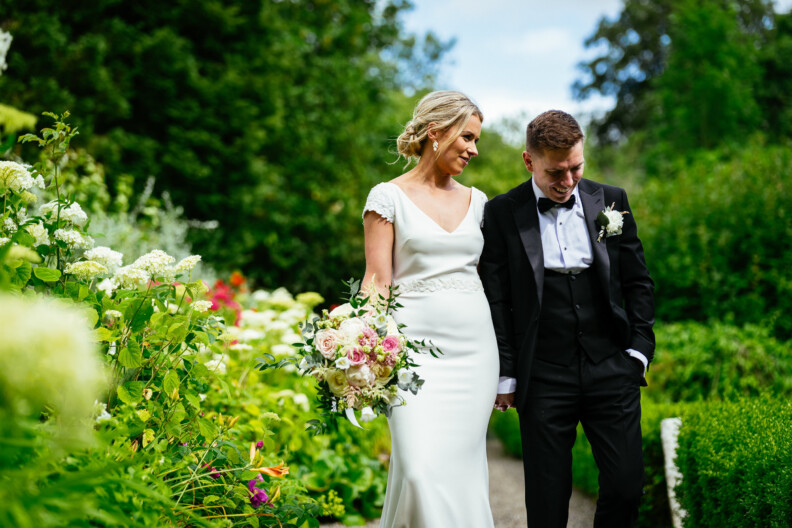 A bride and groom walk hand in hand through a lush garden, surrounded by green foliage and blooming flowers.