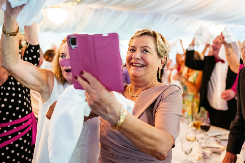 A woman smiling while taking a photo with a tablet at an indoor event, surrounded by people holding up cloths.