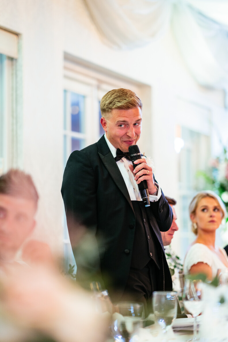 A man in a tuxedo speaks into a microphone at a formal event, with people seated around a table and floral decorations in the background.