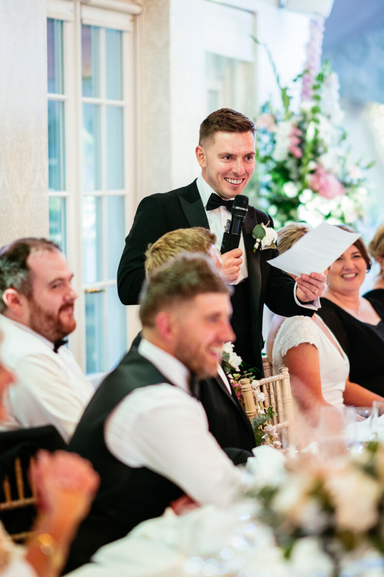 A man in a tuxedo speaks into a microphone while holding a paper, standing at a long table during a formal gathering with people seated nearby.