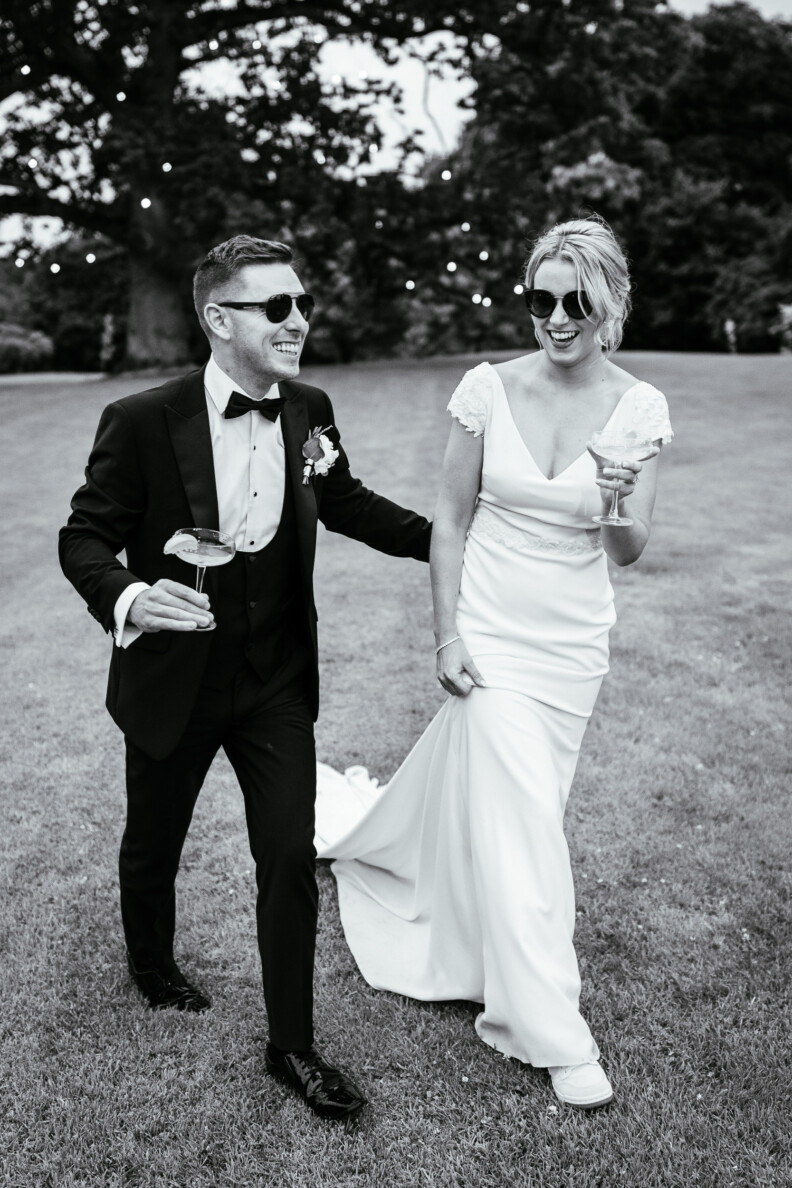 A couple in wedding attire walks on grass, both wearing sunglasses and holding drinks. The man is dressed in a tuxedo, and the woman wears a white wedding dress. They appear joyful.