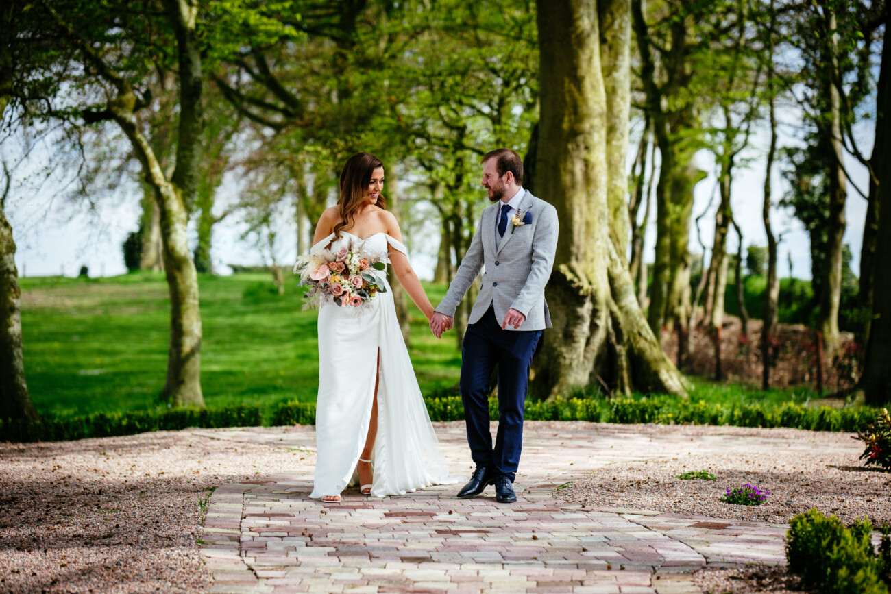 A bride and groom walk hand in hand on a stone path surrounded by trees at their enchanting Darver Castle Wedding, with the bride holding a bouquet.