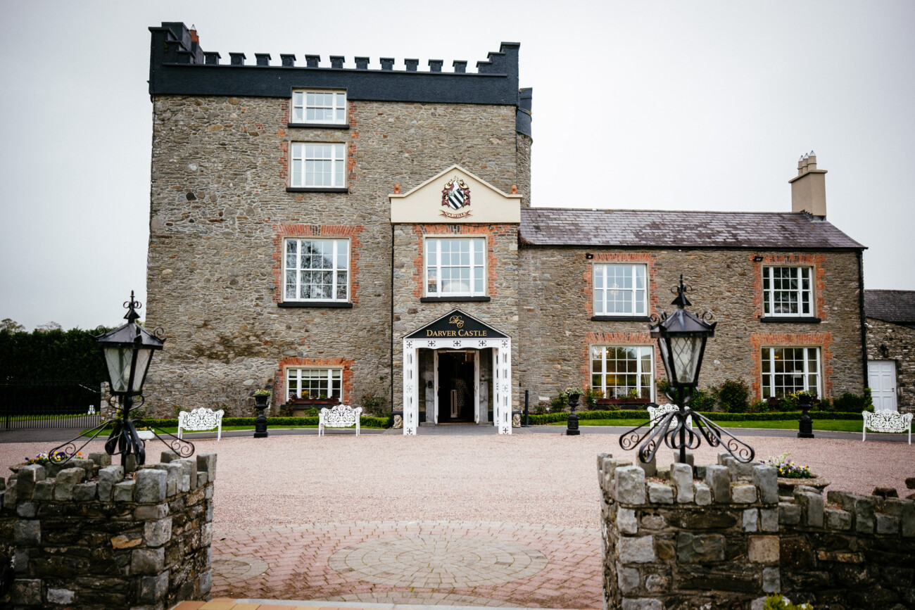 A stone castle with a central tower, surrounded by a cobblestone courtyard and lamps at the entrance. A sign reads "Darver Castle," a perfect setting for an enchanting wedding.