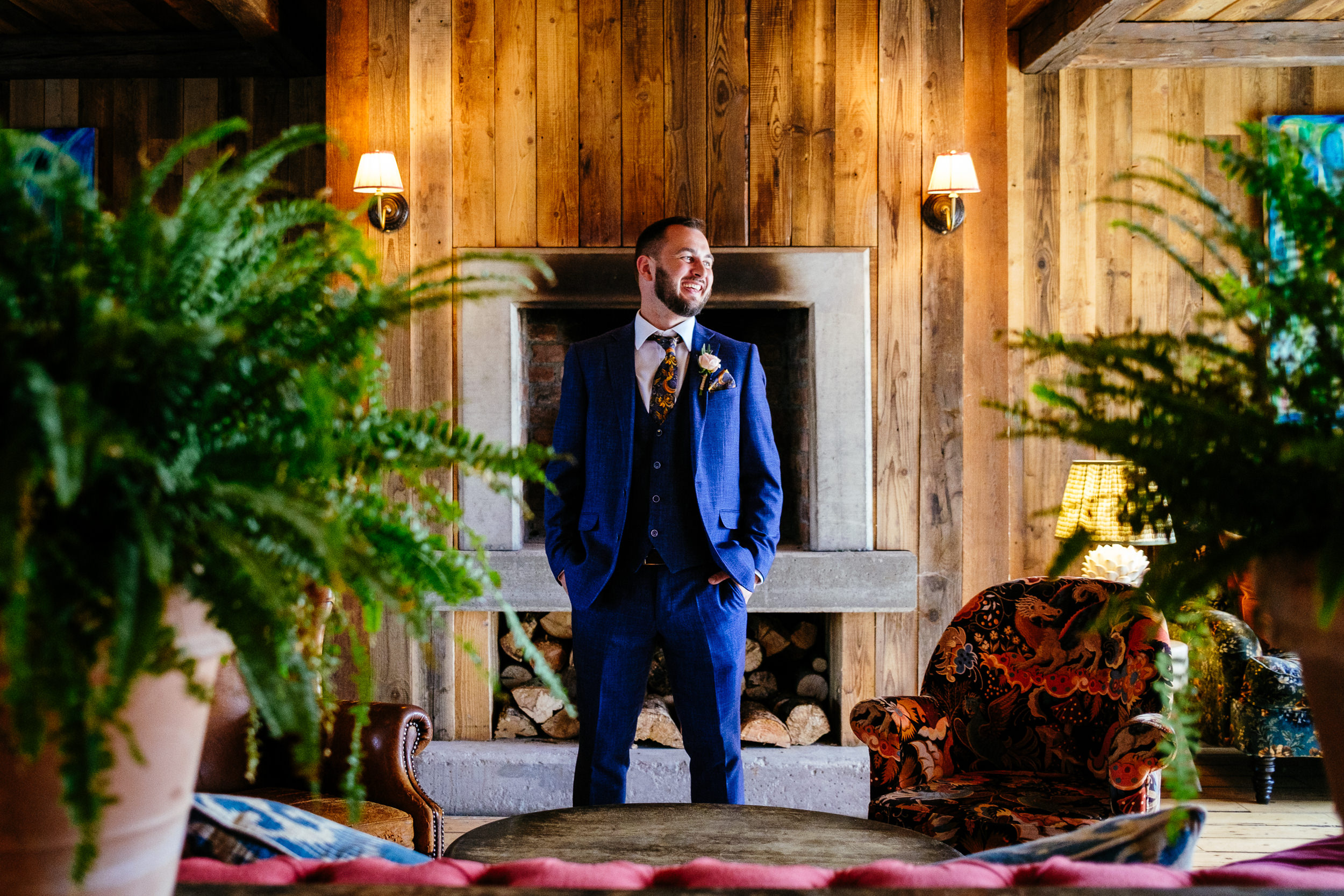A groom in a blue suit stands in a cozy room with wood-paneled walls, reminiscent of a Glasson Lakehouse wedding setting, flanked by two armchairs and lush green plants.