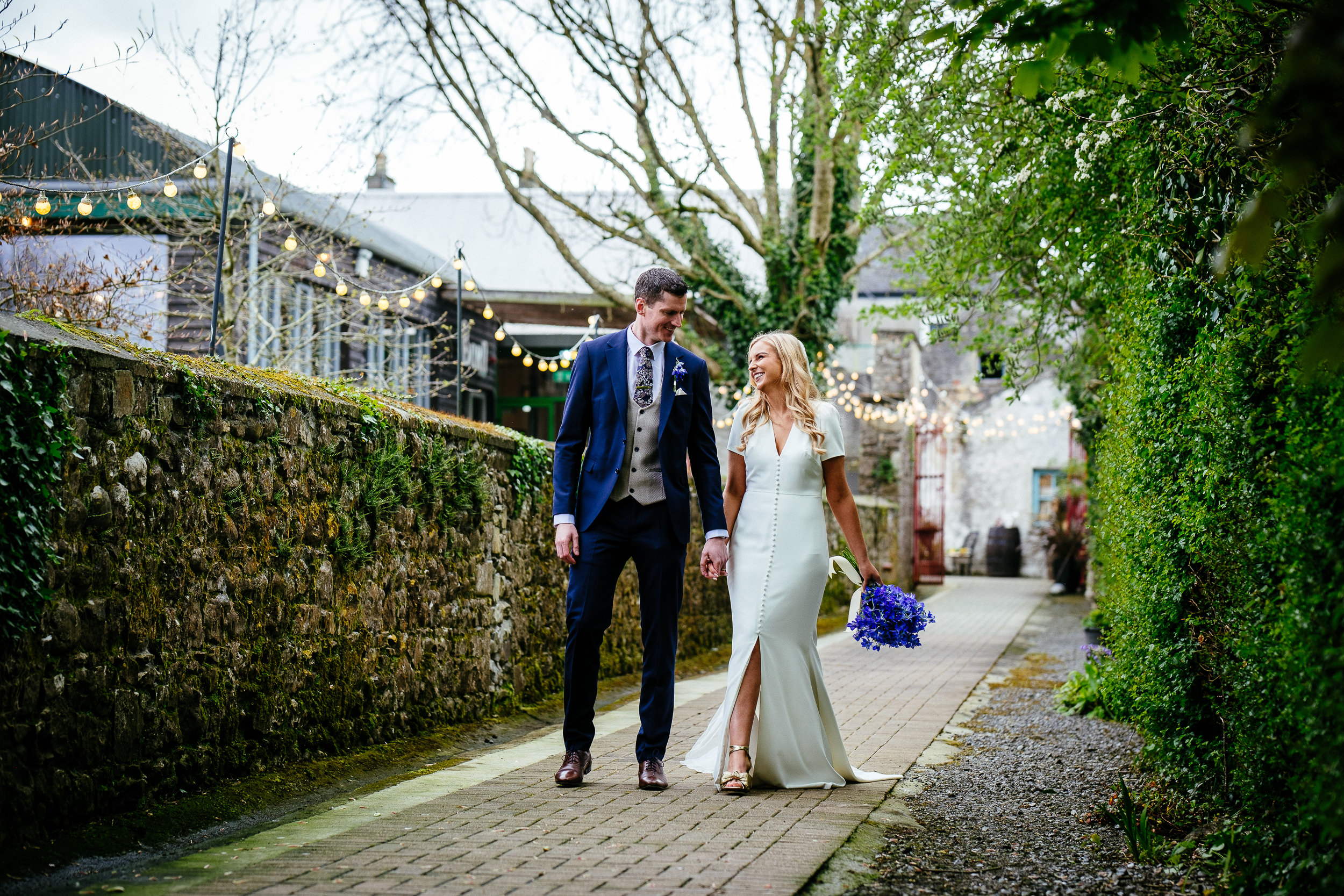 Bride and groom walk hand in hand on a stone path lined with greenery and string lights, with the bride holding a bouquet of purple flowers during their Village Barn Wedding.