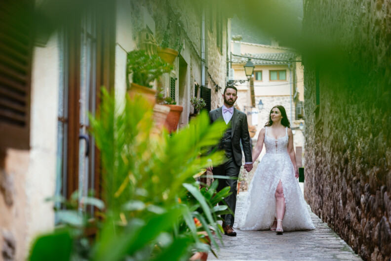 A bride and groom holding hands stroll down a narrow, plant-lined stone alley, creating a perfect scene for destination wedding photography in a rustic setting.