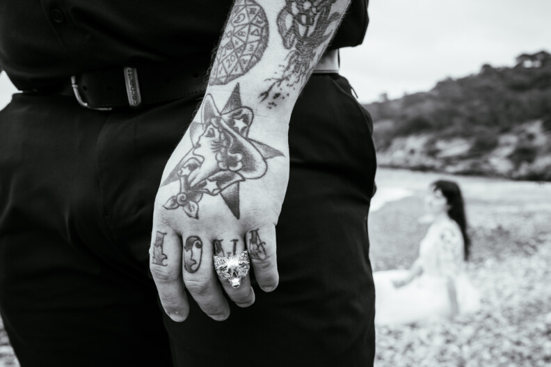 A tattooed hand adorned with rings takes the spotlight, embodying the essence of destination wedding photography as a figure in a white dress sits softly blurred on a rocky beach backdrop.