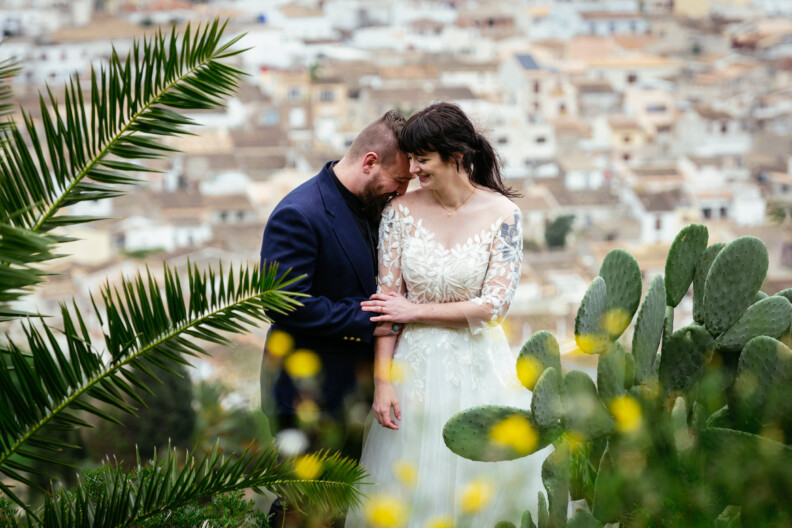 A couple in wedding attire stands close together, enveloped by greenery and yellow flowers, embodying the essence of destination wedding photography with a blurred townscape creating a picturesque backdrop.