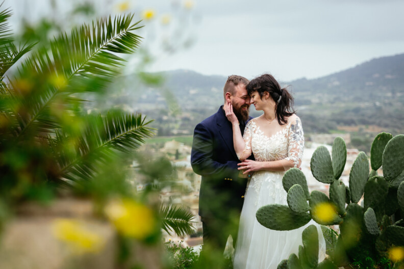 A couple in wedding attire stands close together, surrounded by green foliage and cacti, with distant hills in the background, capturing the essence of destination wedding photography.