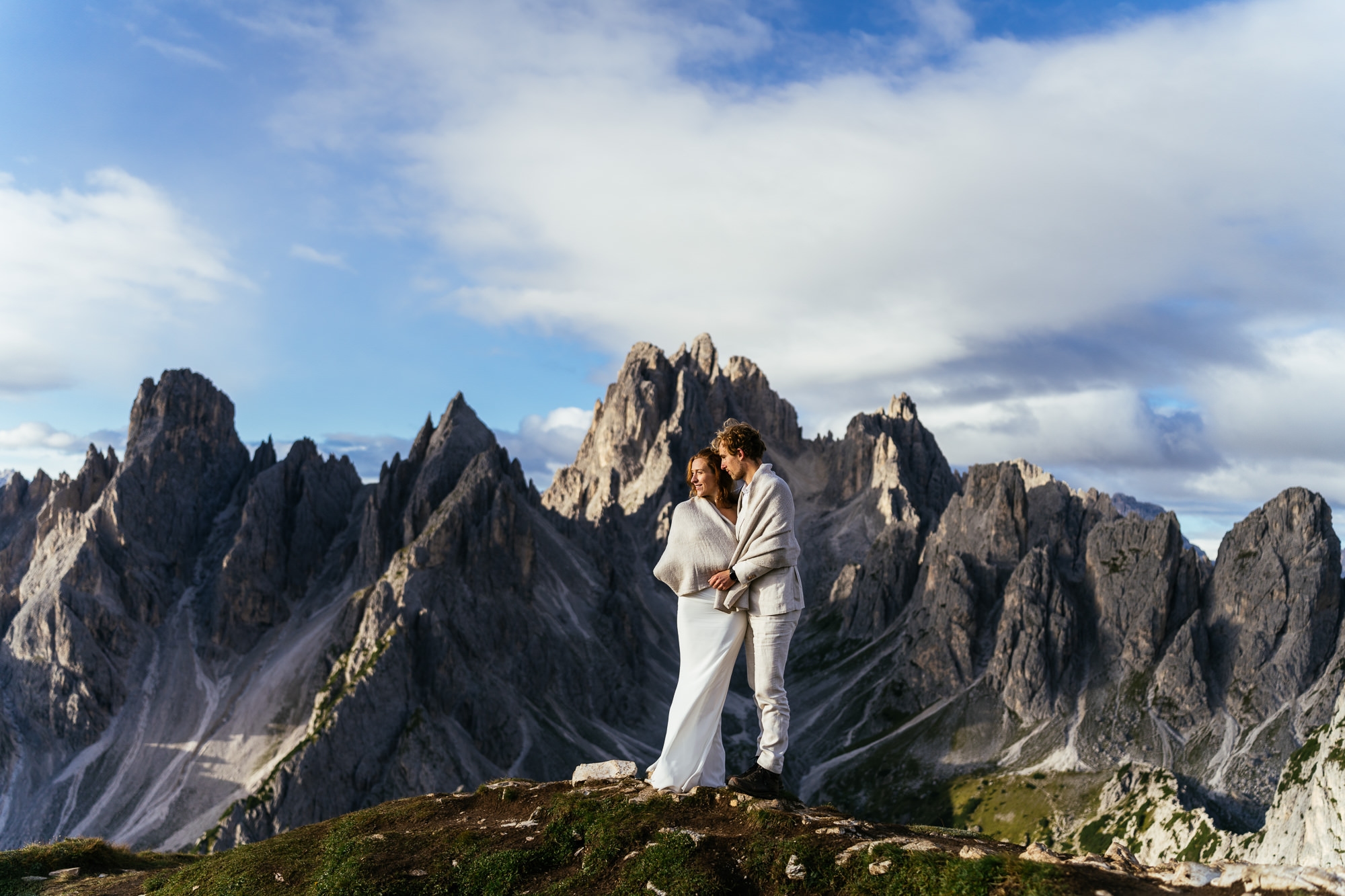 A bride and groom dressed in white stands close together on a grassy hilltop with the rugged, rocky Dolomites in the background under a partly cloudy sky at sunrise during their Bespoke Elopements in Europe.