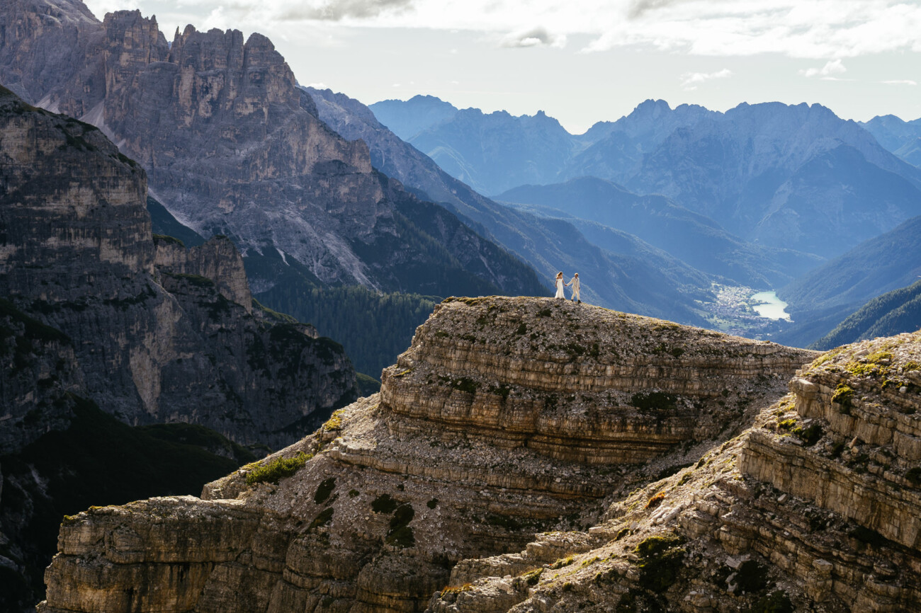 Two people stand on a rocky mountain ledge with dramatic peaks and a distant lake in the background under a partly cloudy sky.