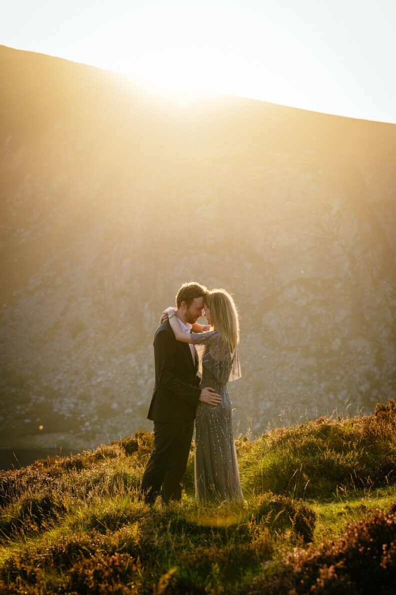 A couple stands embracing on grassy terrain with mountains in the background, illuminated by sunlight from above—a perfect Tulfarris Hotel Wedding moment.