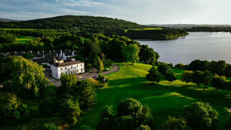 Aerial view of a large the manor, at Tulfarris Hotel Wedding, surrounded by trees and green lawns near a lake, with hills visible in the background.