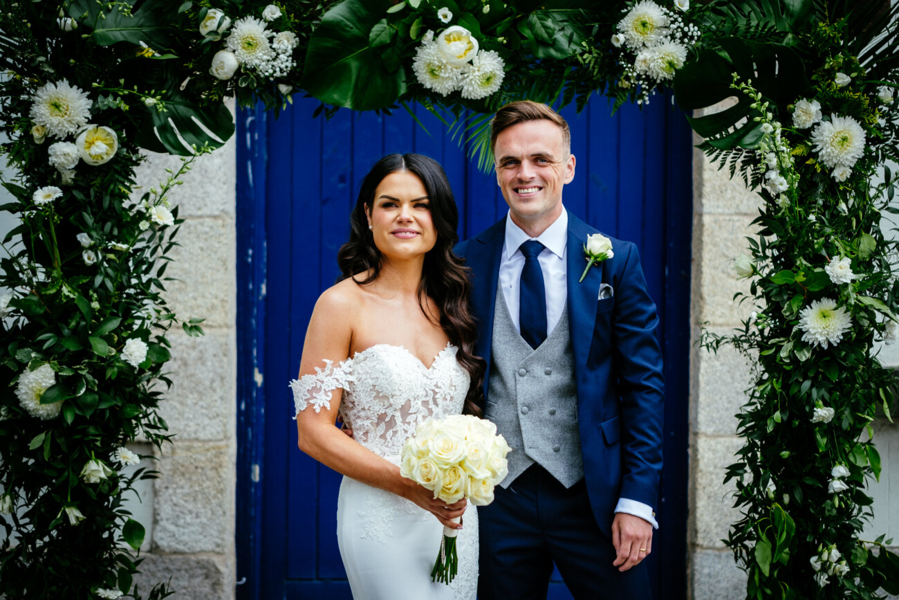 Tara and Darren’s Unforgettable Wedding at Bellingham Castle 86 A bride and groom stand under a flower arch, posing for a wedding photo during their elegant wedding at Bellingham Castle. The bride holds a bouquet of white roses; both are smiling in front of a blue door.