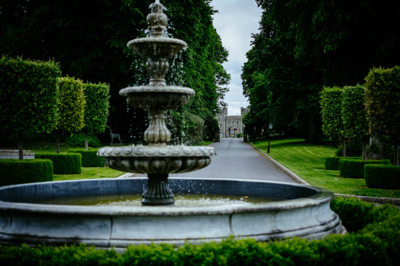 Tara and Darren’s Unforgettable Wedding at Bellingham Castle 87 A tiered stone fountain stands in the foreground of a manicured garden with a tree-lined path leading to a large stone building in the distance, creating an enchanting setting perfect for a wedding at Bellingham Castle.
