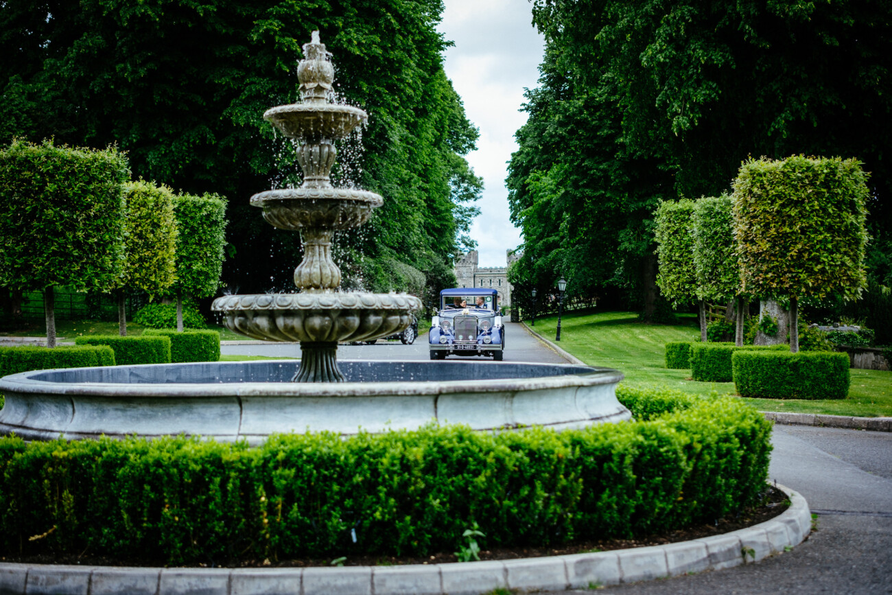 Tara and Darren’s Unforgettable Wedding at Bellingham Castle 88 A stone fountain with water flowing is centred in a manicured garden; behind it, a vintage car drives down the tree-lined road, evoking the timeless elegance of a wedding at Bellingham Castle.