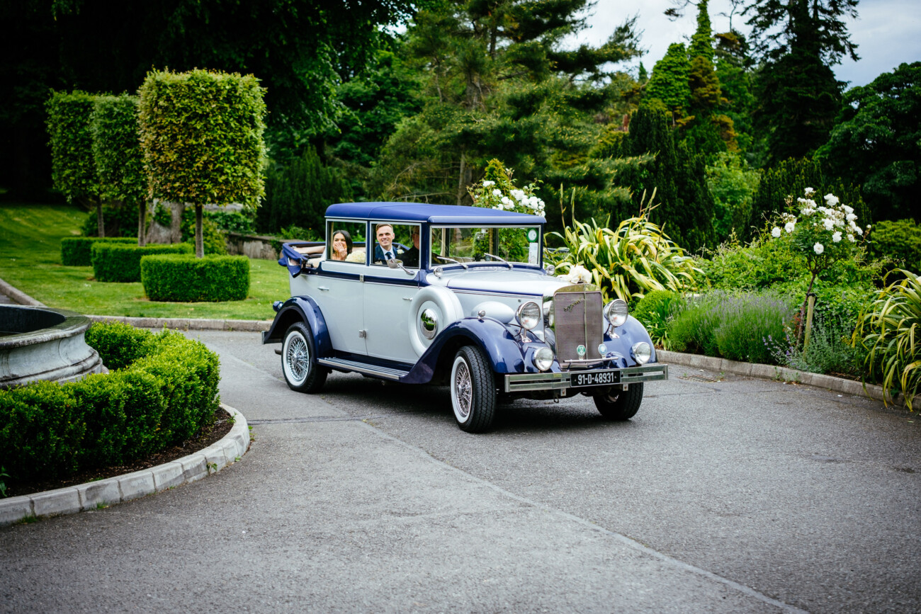 Tara and Darren’s Unforgettable Wedding at Bellingham Castle 89 A vintage car with two people inside drives along a curved drive surrounded by manicured greenery and trees, setting a romantic scene reminiscent of a wedding at Bellingham Castle.