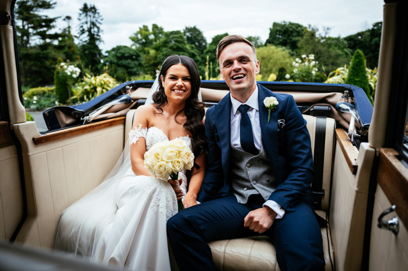 Tara and Darren’s Unforgettable Wedding at Bellingham Castle 90 Bride in a white dress holding a bouquet and groom in a suit sit together in the back seat of a vintage car, smiling at the camera after their beautiful wedding at Bellingham Castle.