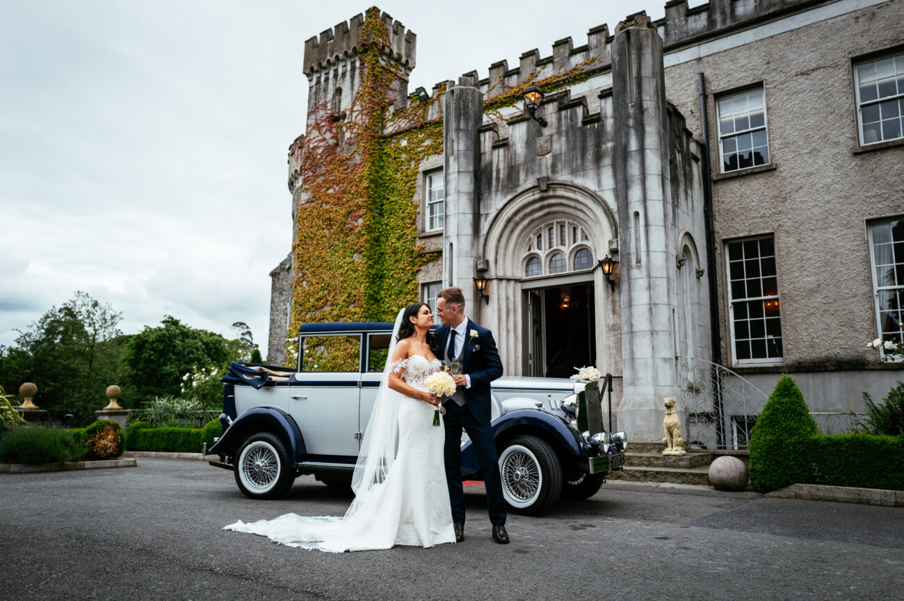 Tara and Darren’s Unforgettable Wedding at Bellingham Castle 91 Bride and groom pose in front of a vintage car outside a stone building covered in greenery and ivy at their elegant wedding at Bellingham Castle, both dressed in formal wedding attire.