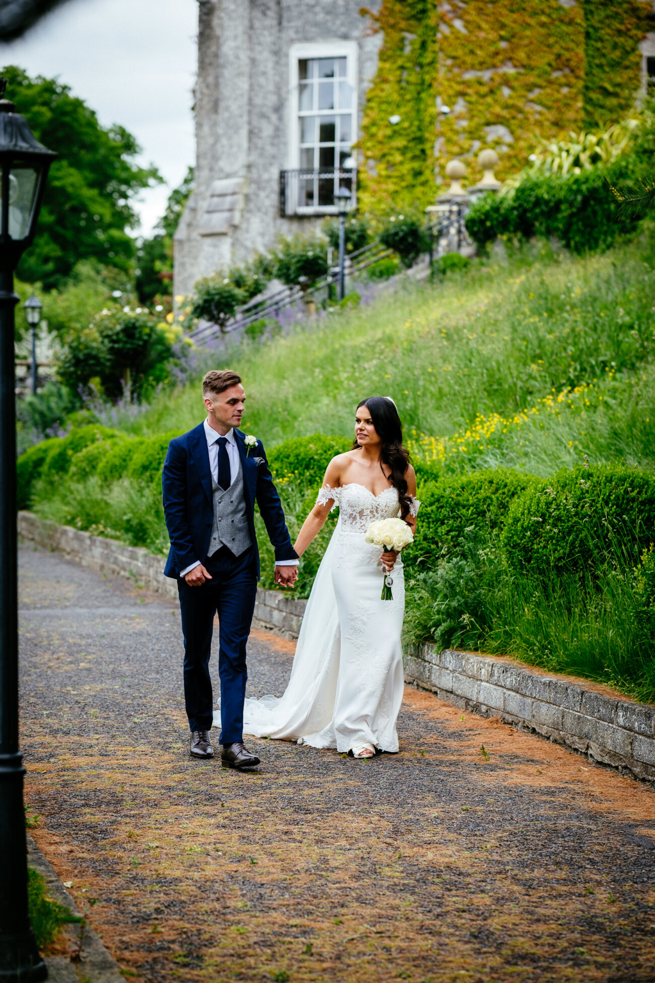 Tara and Darren’s Unforgettable Wedding at Bellingham Castle 92 A bride and groom walk hand in hand on a garden path beside a stone building covered in ivy, capturing the romance of a wedding at Bellingham Castle.