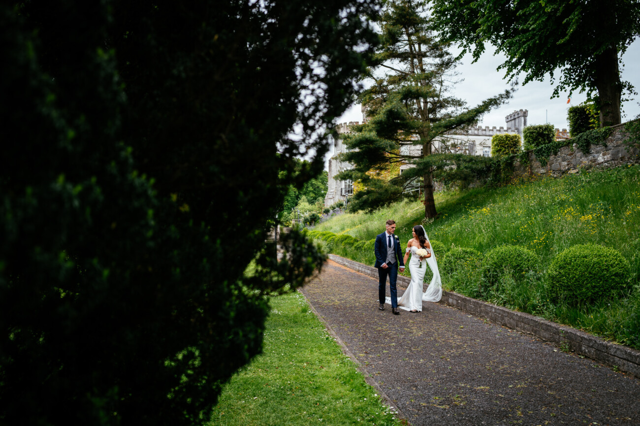 Tara and Darren’s Unforgettable Wedding at Bellingham Castle 93 A bride and groom walk together along a path in a park-like setting, surrounded by trees, grass, and stone walls on a cloudy day during their wedding at Bellingham Castle.