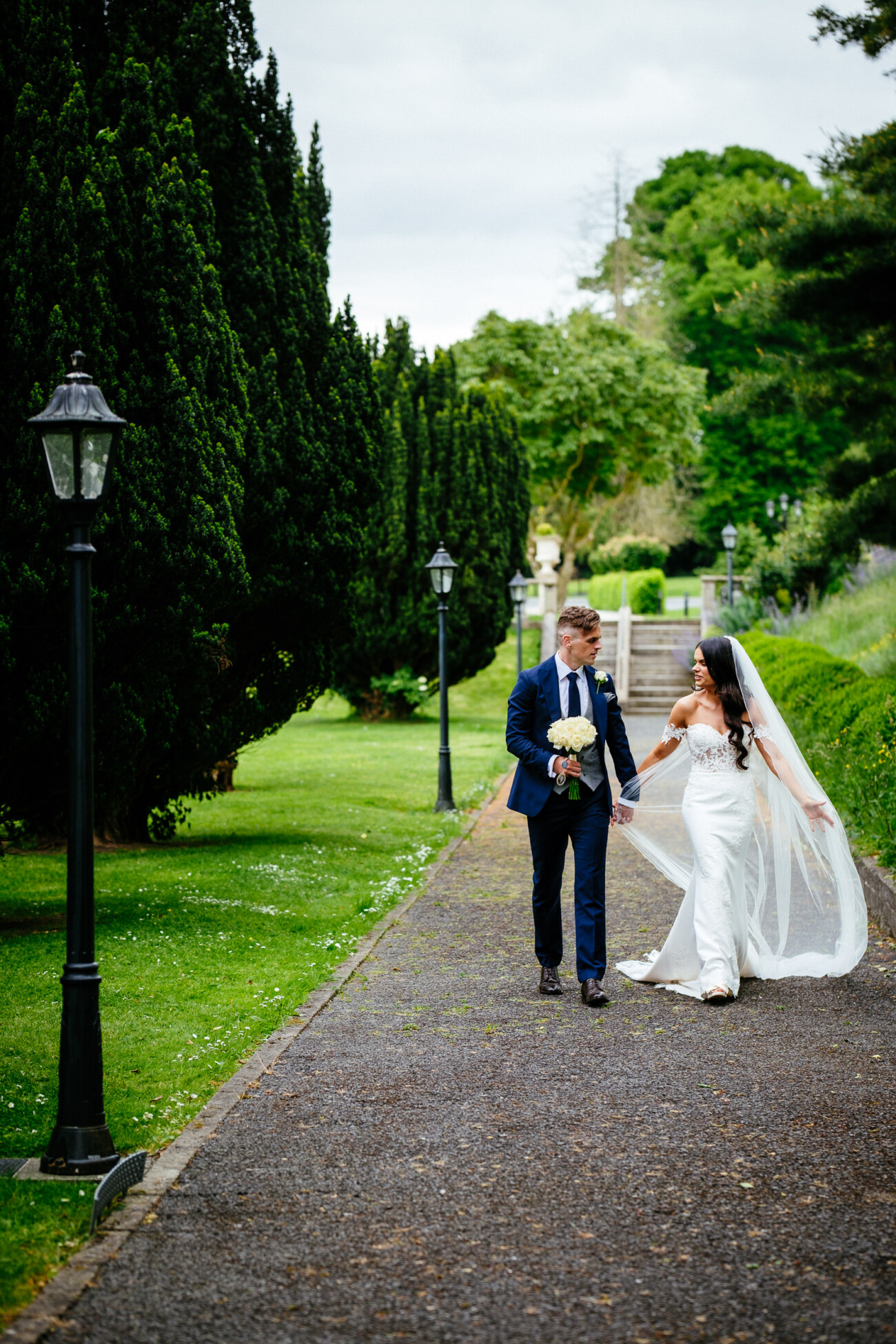 Tara and Darren’s Unforgettable Wedding at Bellingham Castle 94 A bride and groom walk together on a tree-lined path in a park during their wedding at Bellingham Castle, with the bride holding her bouquet and veil whilst smiling at the groom.