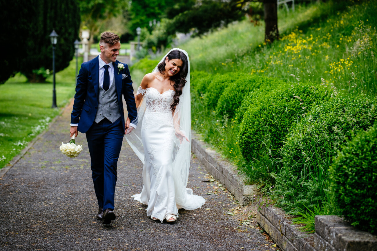 Tara and Darren’s Unforgettable Wedding at Bellingham Castle 95 A bride in a white dress and veil walks hand in hand with a groom in a navy suit holding flowers on a garden path during their romantic wedding at Bellingham Castle.