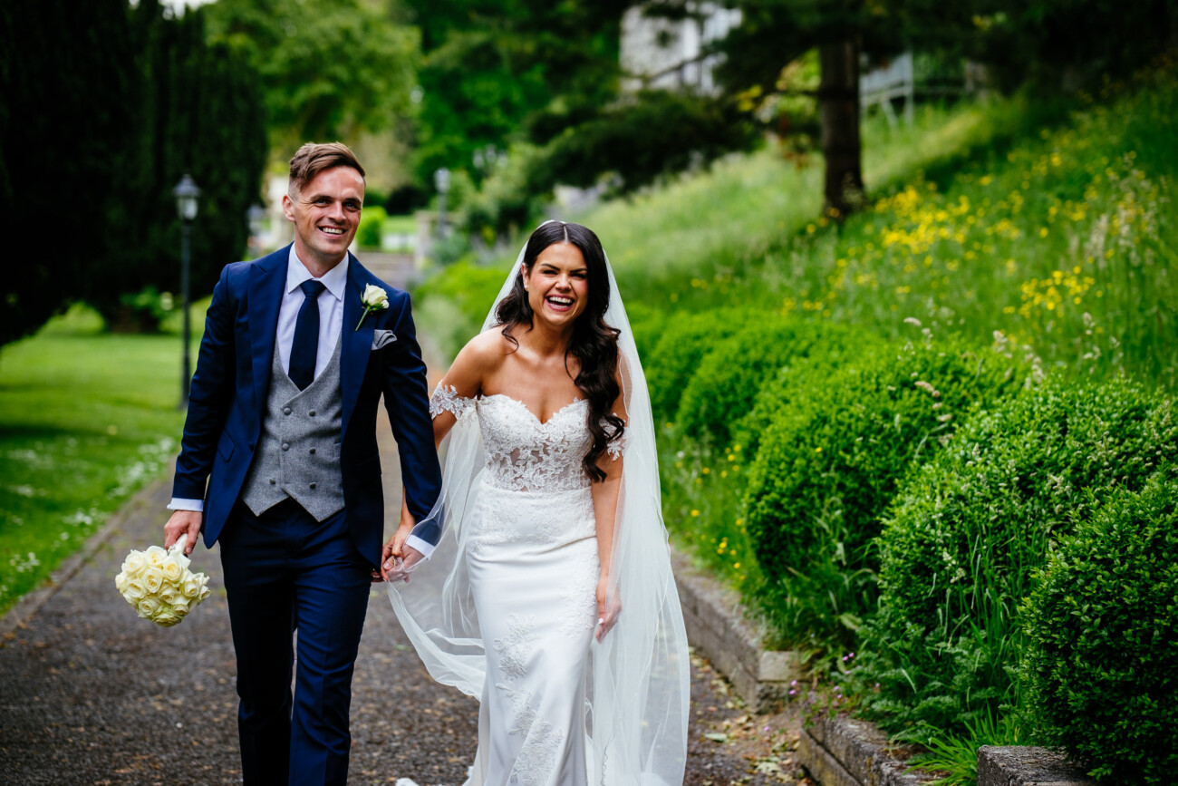 Tara and Darren’s Unforgettable Wedding at Bellingham Castle 96 A bride and groom walk outside on a path, holding hands and smiling during their wedding at Bellingham Castle. The groom holds a bouquet of white flowers, as greenery and trees surround them.