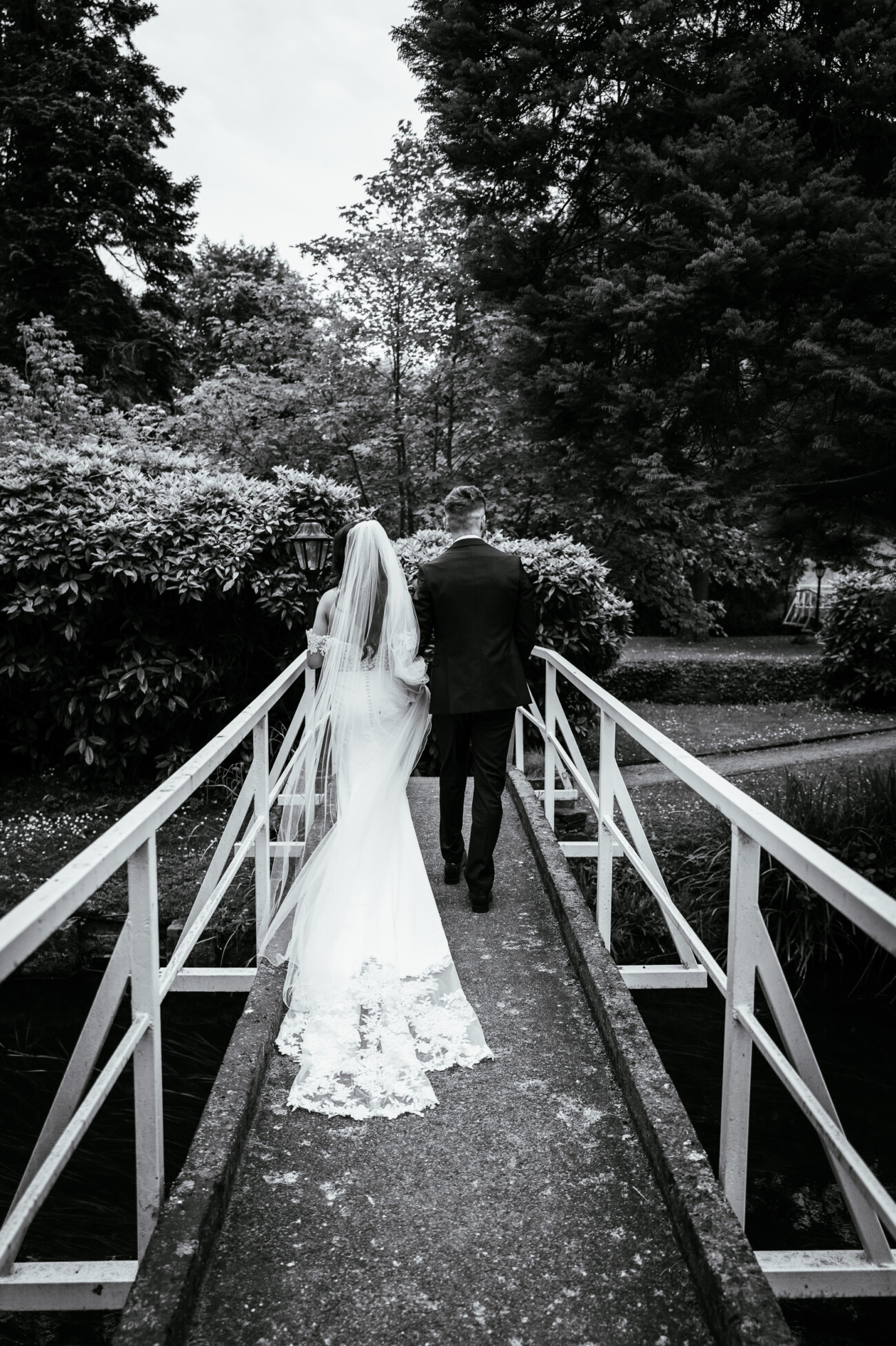 Tara and Darren’s Unforgettable Wedding at Bellingham Castle 97 A bride and groom walk together across a narrow bridge in a garden, photographed from behind in black and white during their wedding at Bellingham Castle.