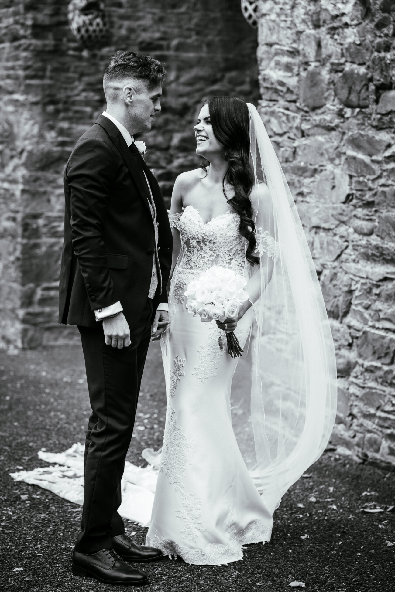 Tara and Darren’s Unforgettable Wedding at Bellingham Castle 98 A bride in a white dress and veil holds a bouquet while standing and smiling at a groom in a suit outdoors near a stone wall, beautifully capturing the romance of their wedding at Bellingham Castle.