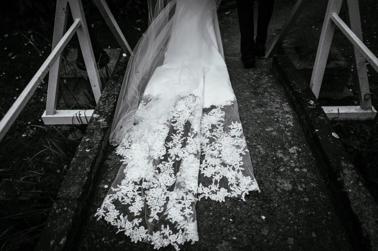 Tara and Darren’s Unforgettable Wedding at Bellingham Castle 99 A bride in a white lace dress and veil walks over a small concrete bridge during a wedding at Bellingham Castle, with only the lower part of her gown and someone’s legs visible.
