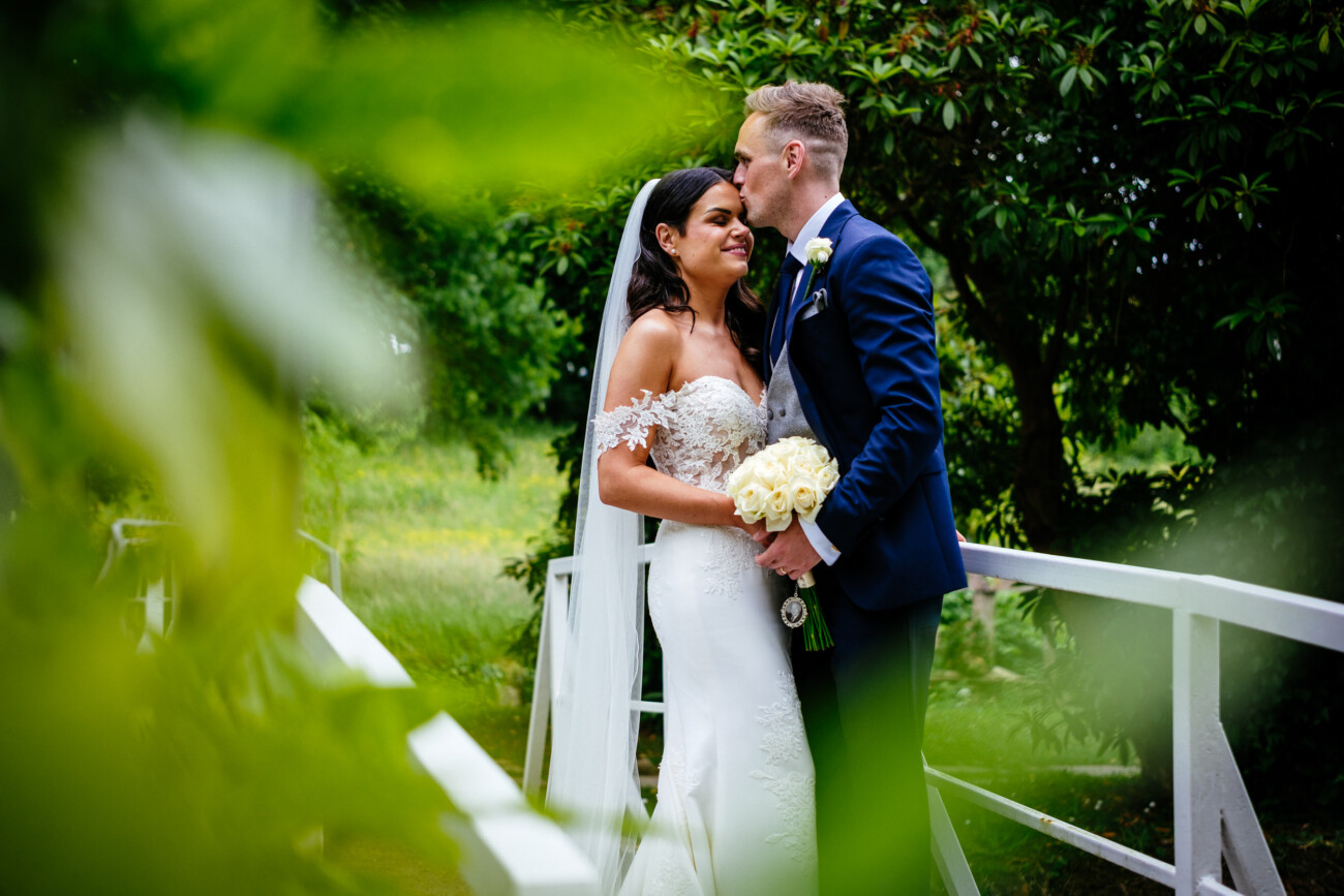 Tara and Darren’s Unforgettable Wedding at Bellingham Castle 100 A groom in a navy suit kisses his bride’s forehead as she holds a bouquet of white roses, standing together on a white bridge surrounded by greenery during their wedding at Bellingham Castle.