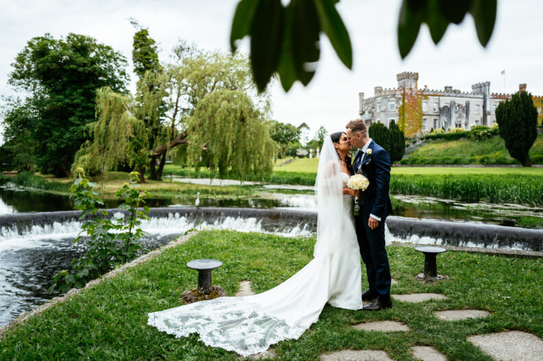 A bride and groom stand together beside a river, with a castle and trees in the background. Celebrating their wedding at Bellingham Castle, the bride wears a long white veil and holds a bouquet whilst the groom kisses her forehead.