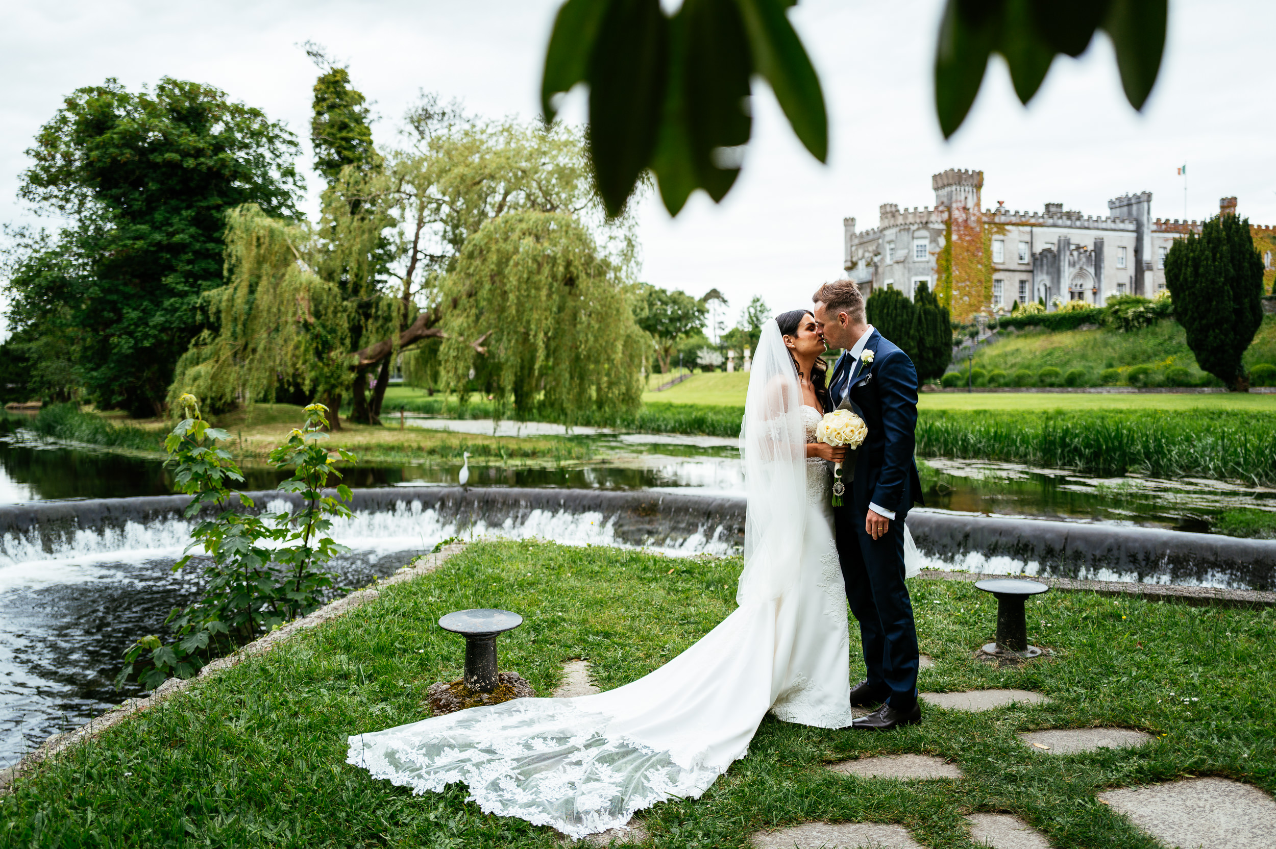 A bride and groom stand together beside a river, with a castle and trees in the background. Celebrating their wedding at Bellingham Castle, the bride wears a long white veil and holds a bouquet whilst the groom kisses her forehead.