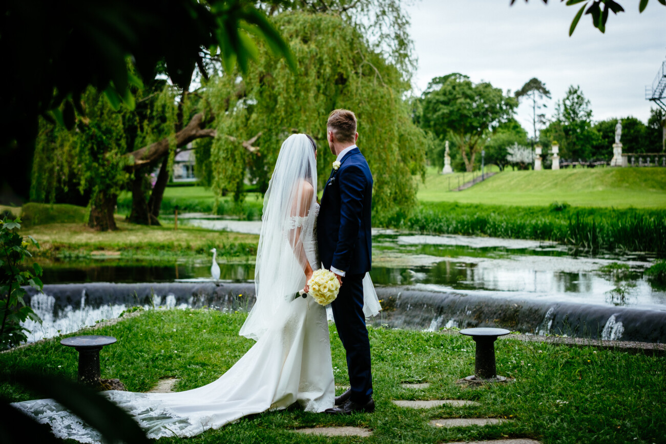 Tara and Darren’s Unforgettable Wedding at Bellingham Castle 102 A bride and groom stand together outdoors by a small waterfall, facing away from the camera, with greenery and trees surrounding them during their romantic wedding at Bellingham Castle.